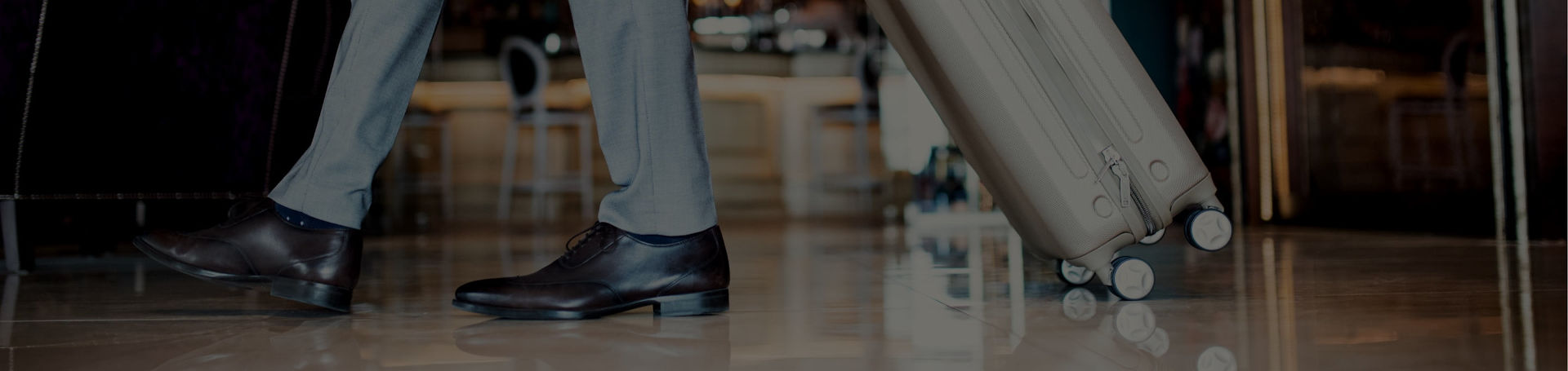 Los pies de una persona con traje y zapatos llevando una maleta con ruedas